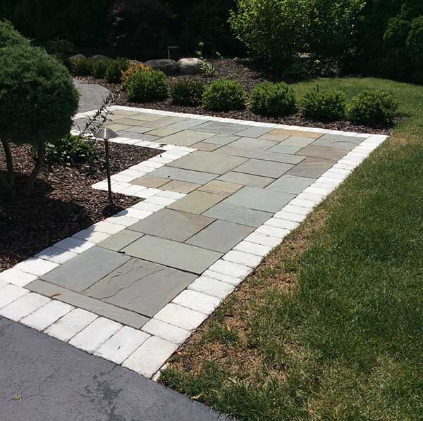 A stone path featuring grey rectangular pavers with a white brick border, curving past mulch beds and a green lawn.
