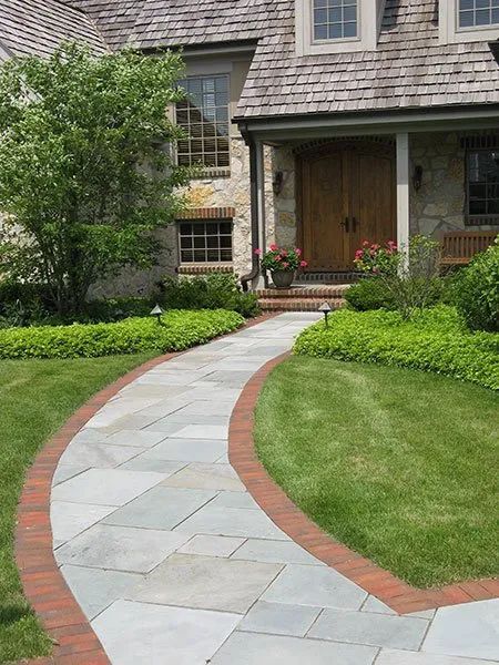 A stone walkway with a red brick border leads through a lush green lawn to the entrance of a stone house.