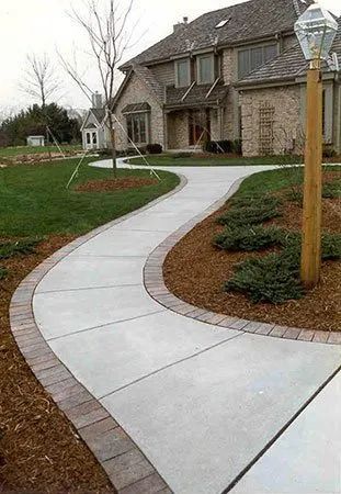 A winding concrete walkway with a decorative brick border leads to the entrance of a large brick suburban house.