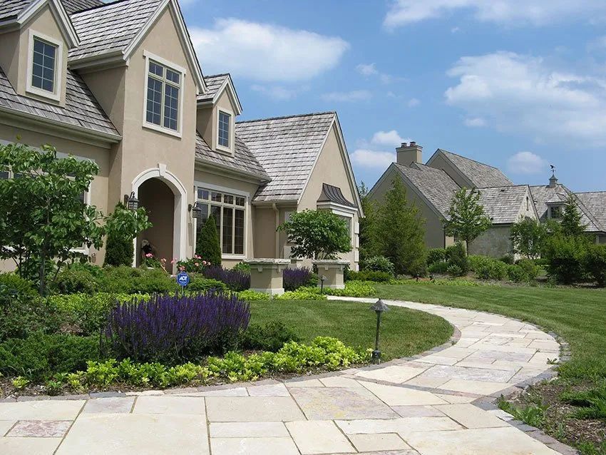 A tan house with a cedar shake roof and stone walkway set in a sunny, manicured suburban lawn with flower beds.