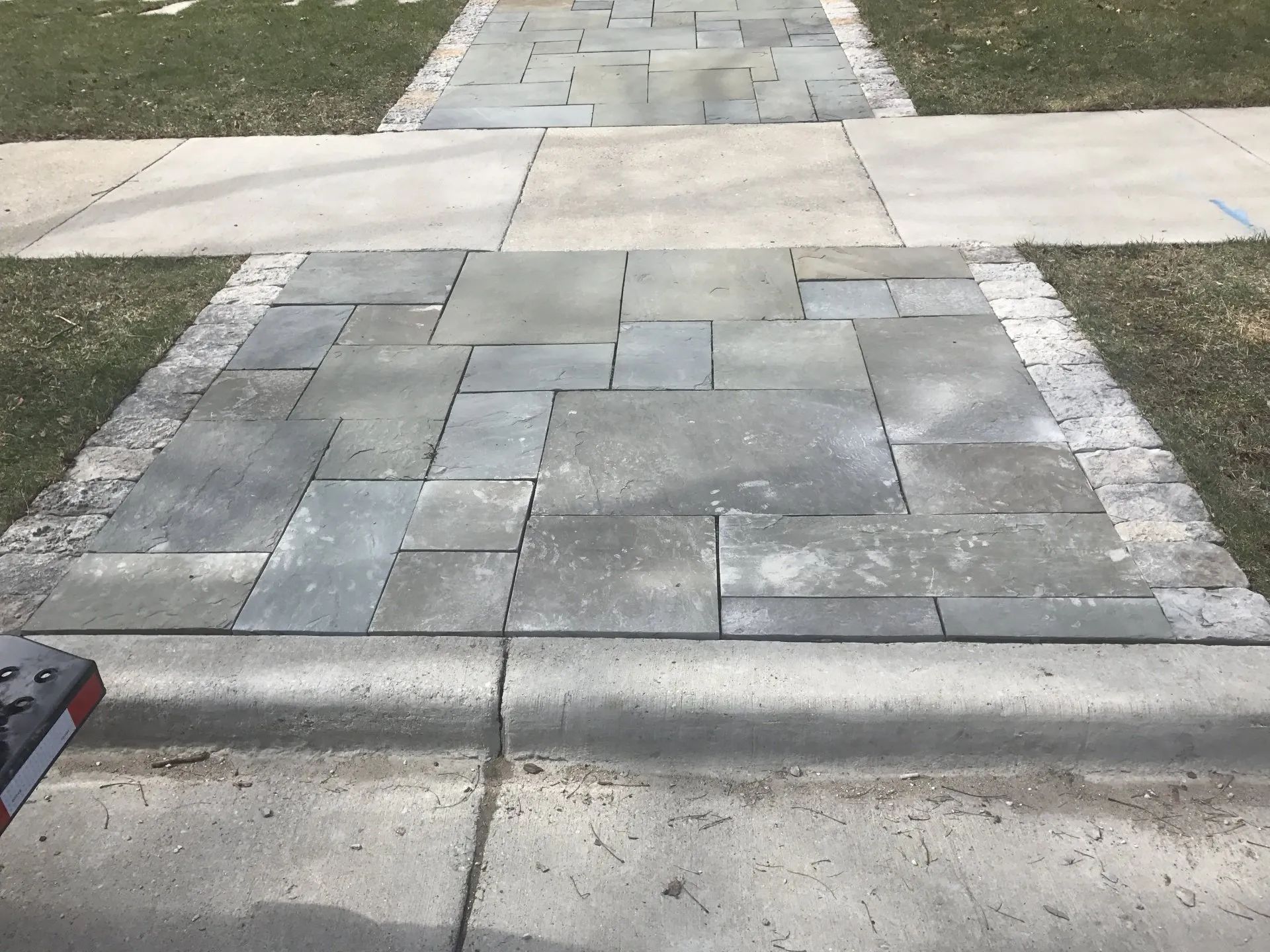 A stone walkway with a decorative border sits between a concrete sidewalk and a curb.