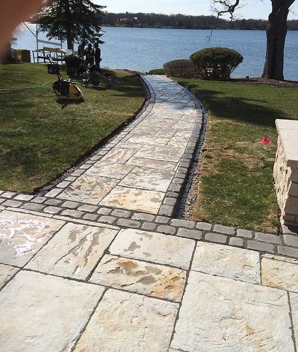A stone walkway with a dark border leads from a paved patio toward a lake on a sunny day.