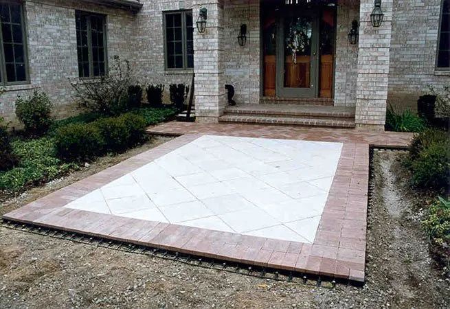 A stone patio with a light-colored central inlay surrounded by a reddish-brown brick border in front of a house entrance.