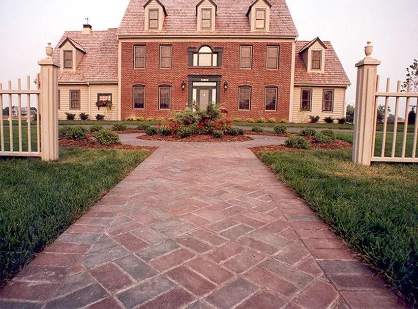 A brick walkway leads to the front entrance of a two-story colonial-style brick house with a gated yard.