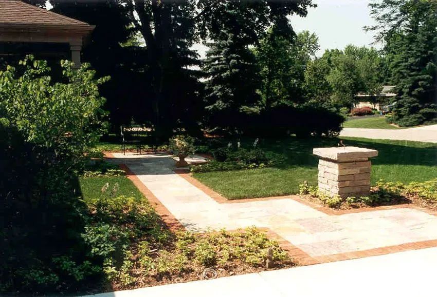A stone paver walkway with brick borders leads through a lush green yard toward a stone pedestal and trees.