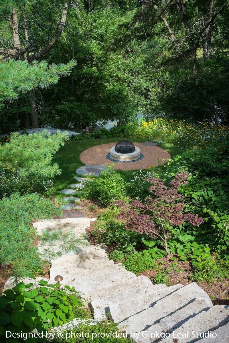 Stone steps lead down through a lush, green garden to a circular fire pit area surrounded by trees and wildflowers.