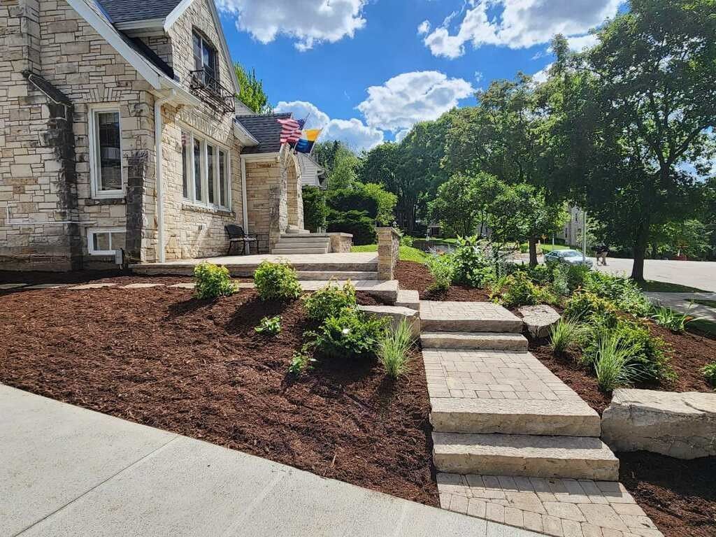 A stone house with landscaped mulch beds, stone stairs, and a path leading to the entrance under a blue sky.