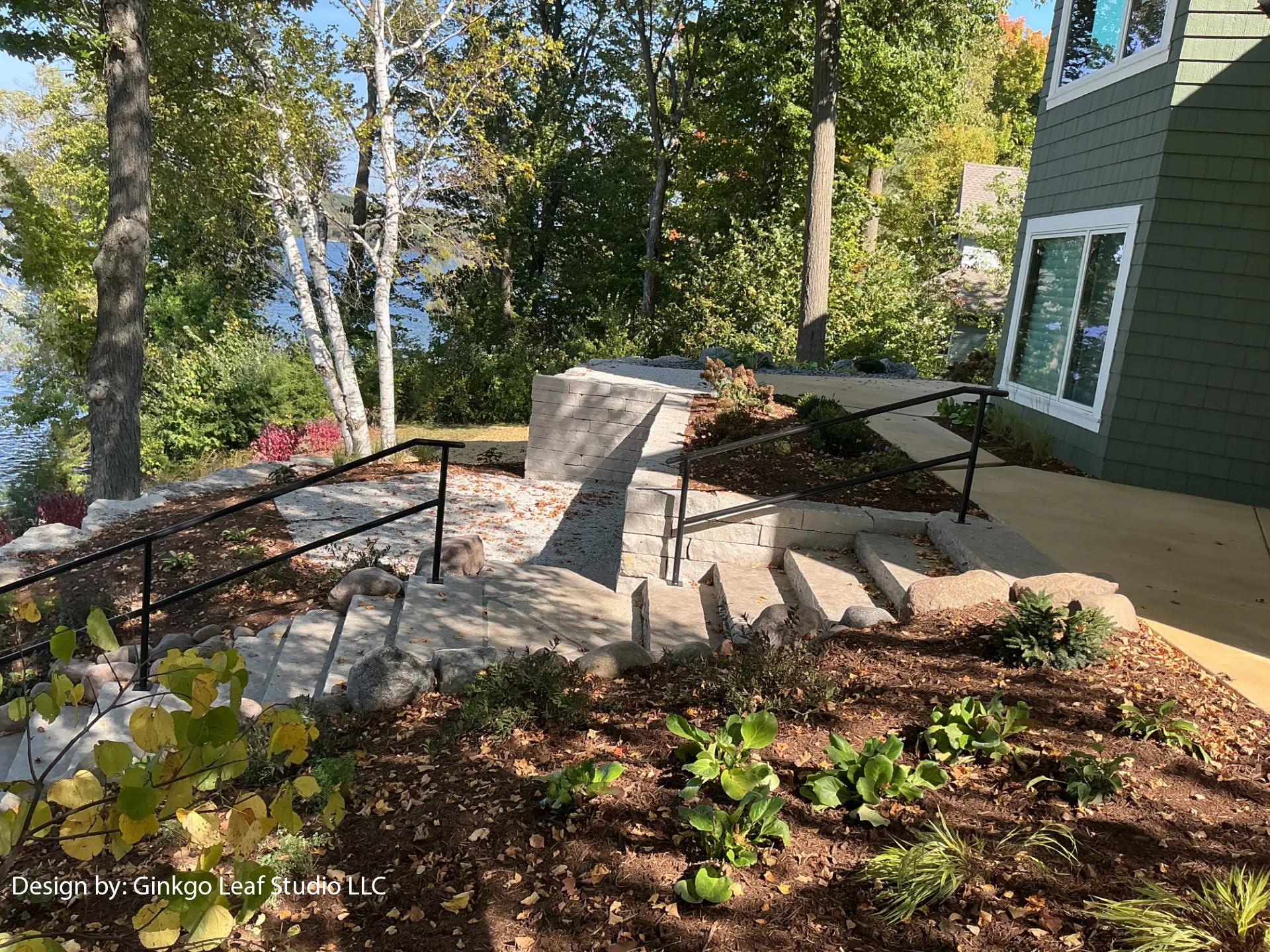 Outdoor stone steps with metal railings lead down a landscaped slope toward a lake near a green house exterior.