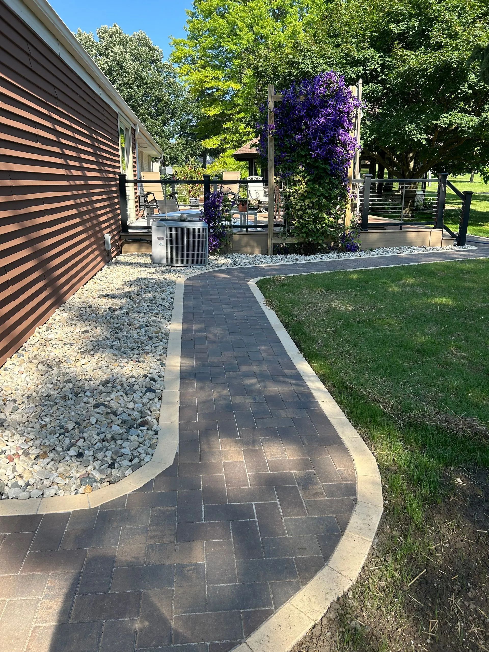 A stone paver walkway with a light-colored border curves alongside a house wall, leading toward a patio with purple flowers.