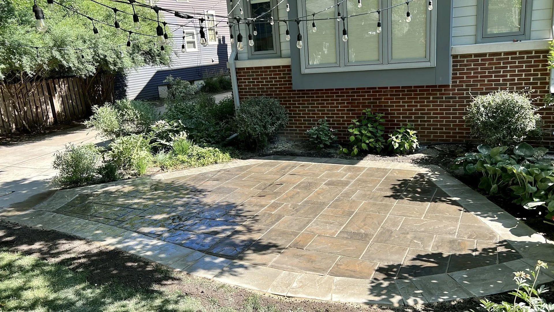 A rectangular, tan-toned paver patio sits in a garden next to a brick house exterior under string lights and trees.