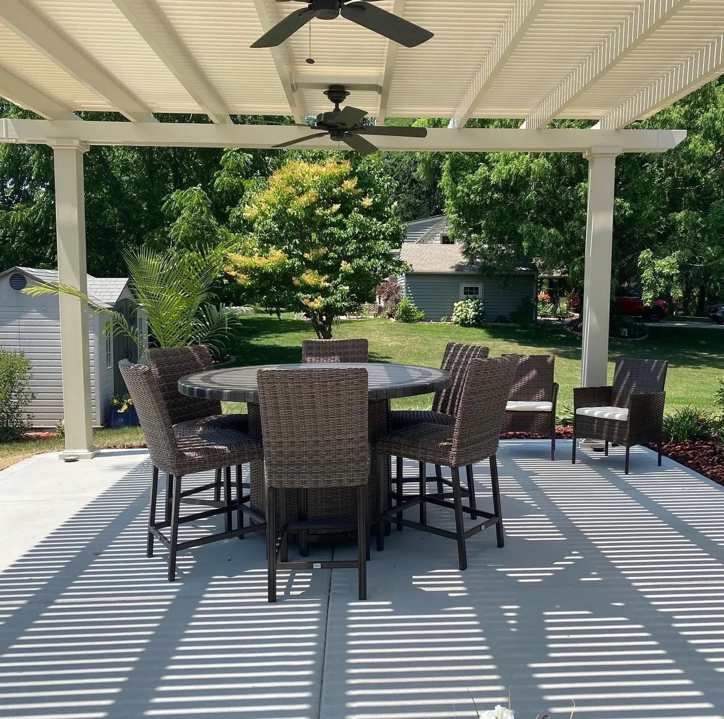 A patio set with a round table and six wicker chairs under a pergola on a concrete deck.