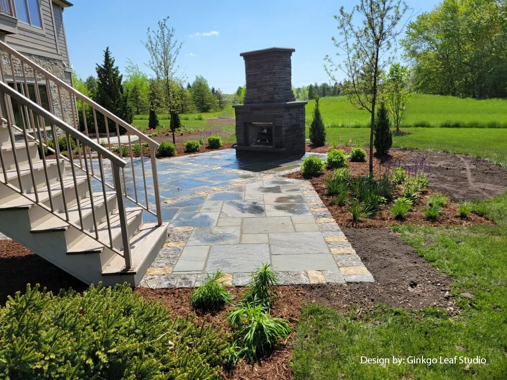 A stone patio with an outdoor fireplace sits next to a residential staircase, surrounded by lawn and new garden plantings.