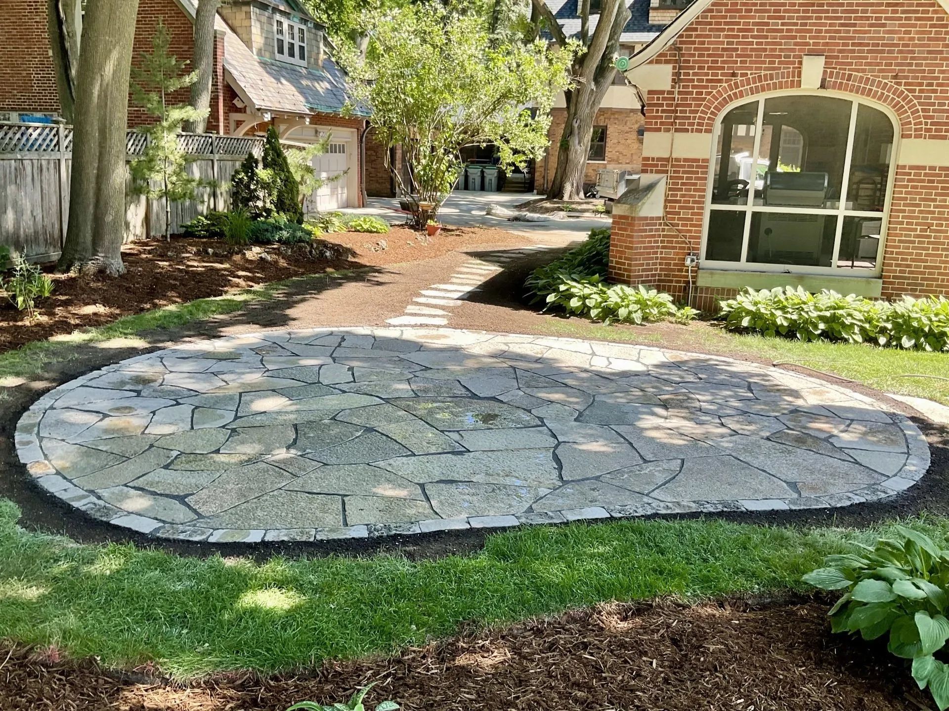 A flagstone patio with a curved stone border set in a backyard between two brick houses on a sunny day.