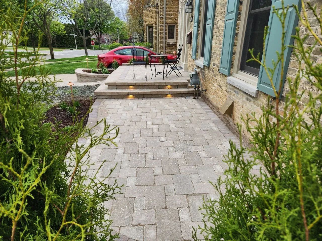 A stone walkway leads to a raised patio with steps and landscape lighting in front of a stone house with blue shutters.