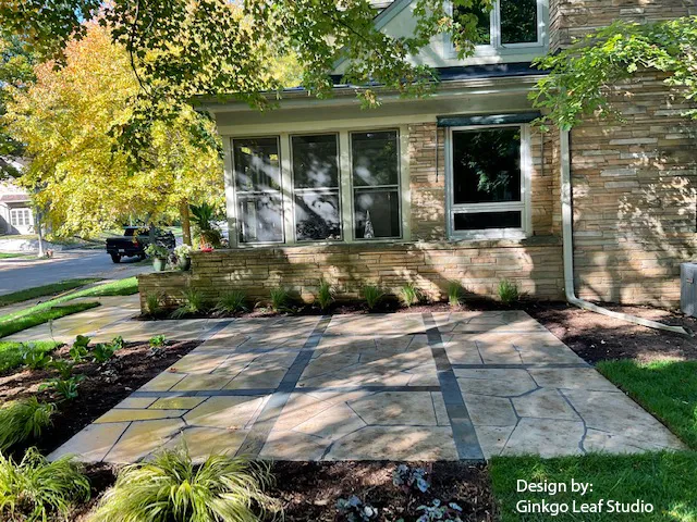 A stamped concrete patio featuring flagstone-patterned tiles accented by dark geometric borders in front of a stone house.