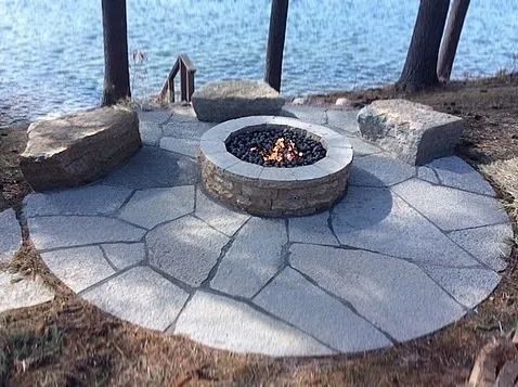 A circular stone patio features a fire pit with a lit fire, surrounded by three large natural stone benches by a lake.