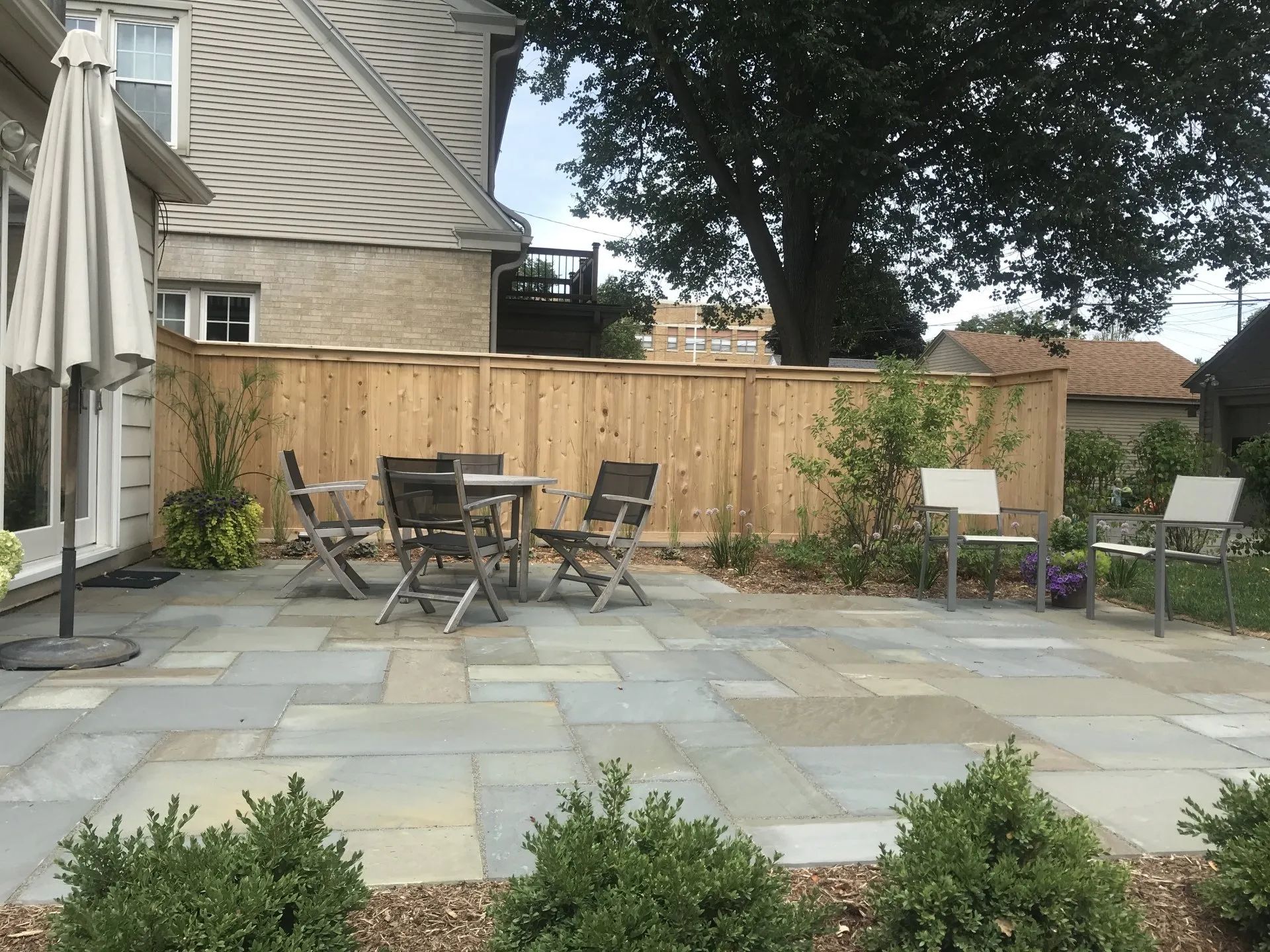 A backyard patio featuring a stone floor, outdoor dining set, two chairs, a large umbrella, and a wooden privacy fence.