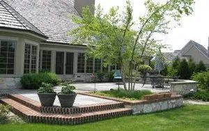 A stone patio with brick-edged steps and potted plants sits behind a house, with a small tree and lawn in the foreground.