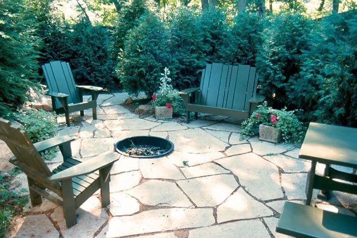 A patio with a stone fire pit, Adirondack-style chairs, and a table, surrounded by lush green trees and potted plants.