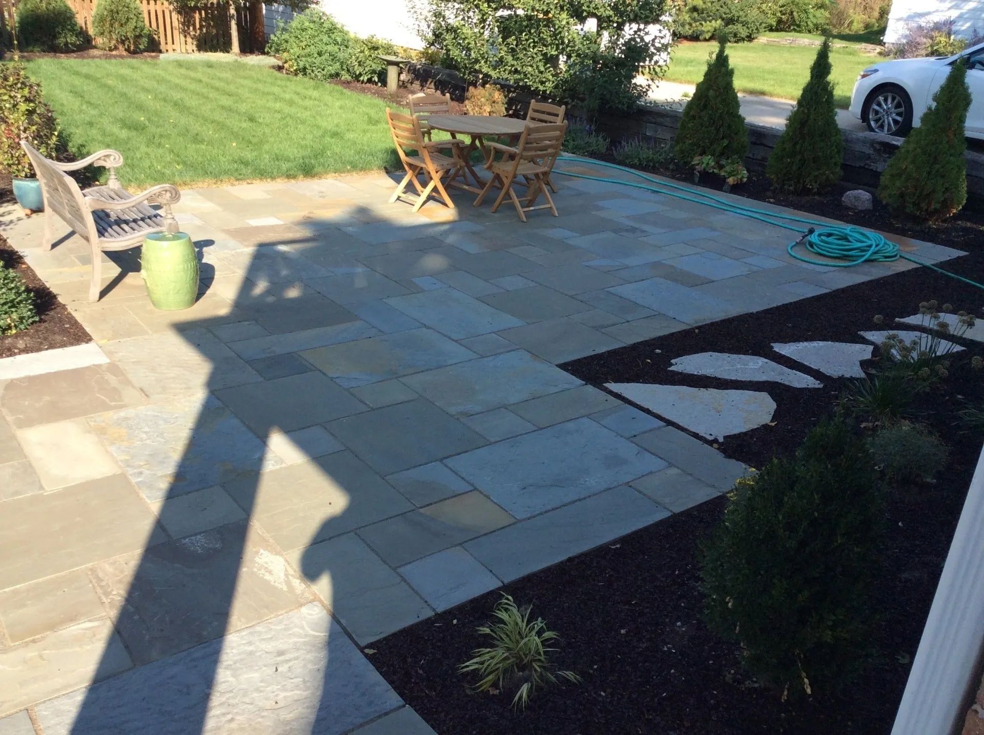 A stone patio with a dining set and bench, bordered by mulch beds, small evergreen shrubs, and a lawn.