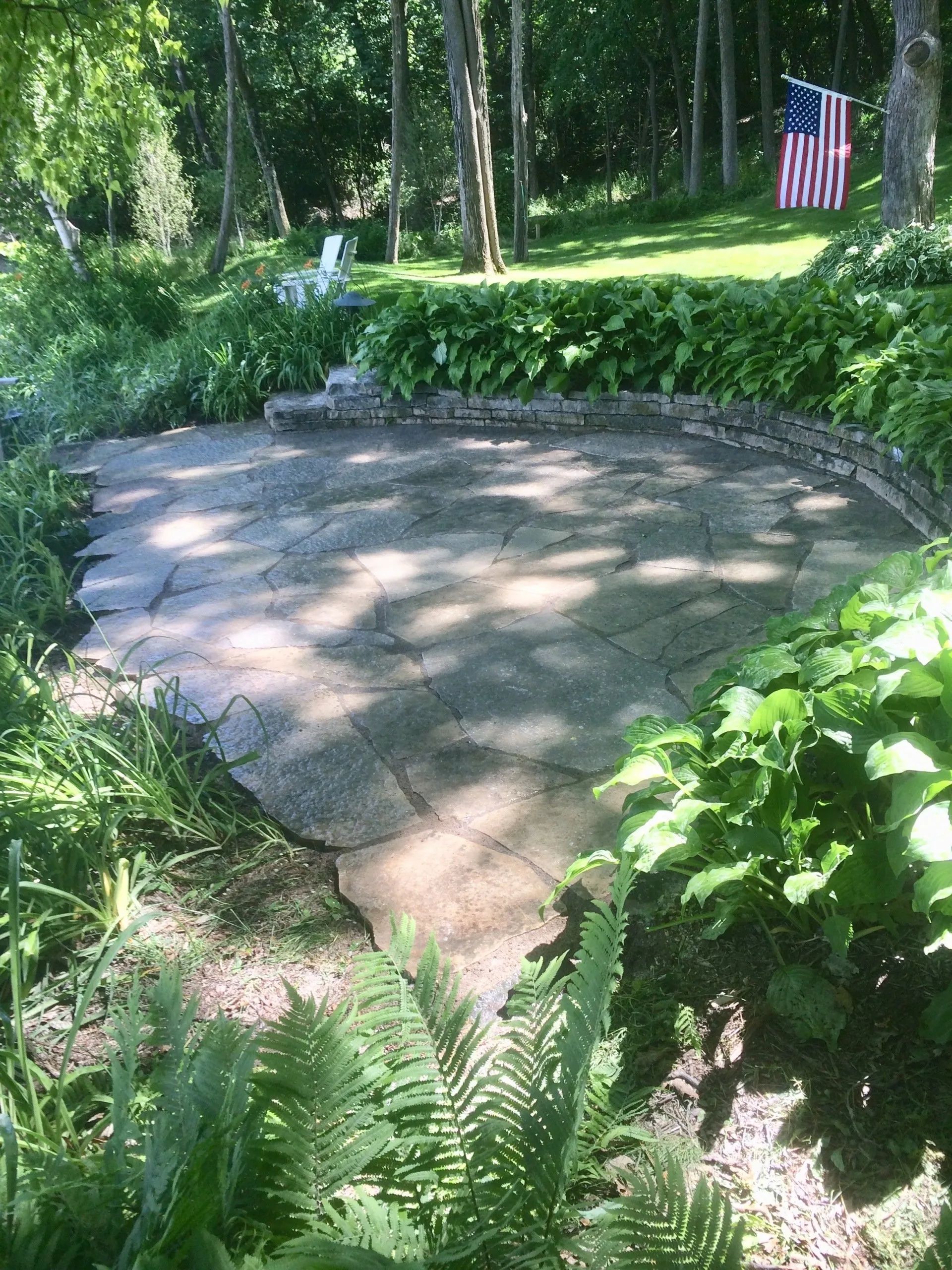 A stone patio surrounded by lush green foliage and trees, with an American flag flying in the background.