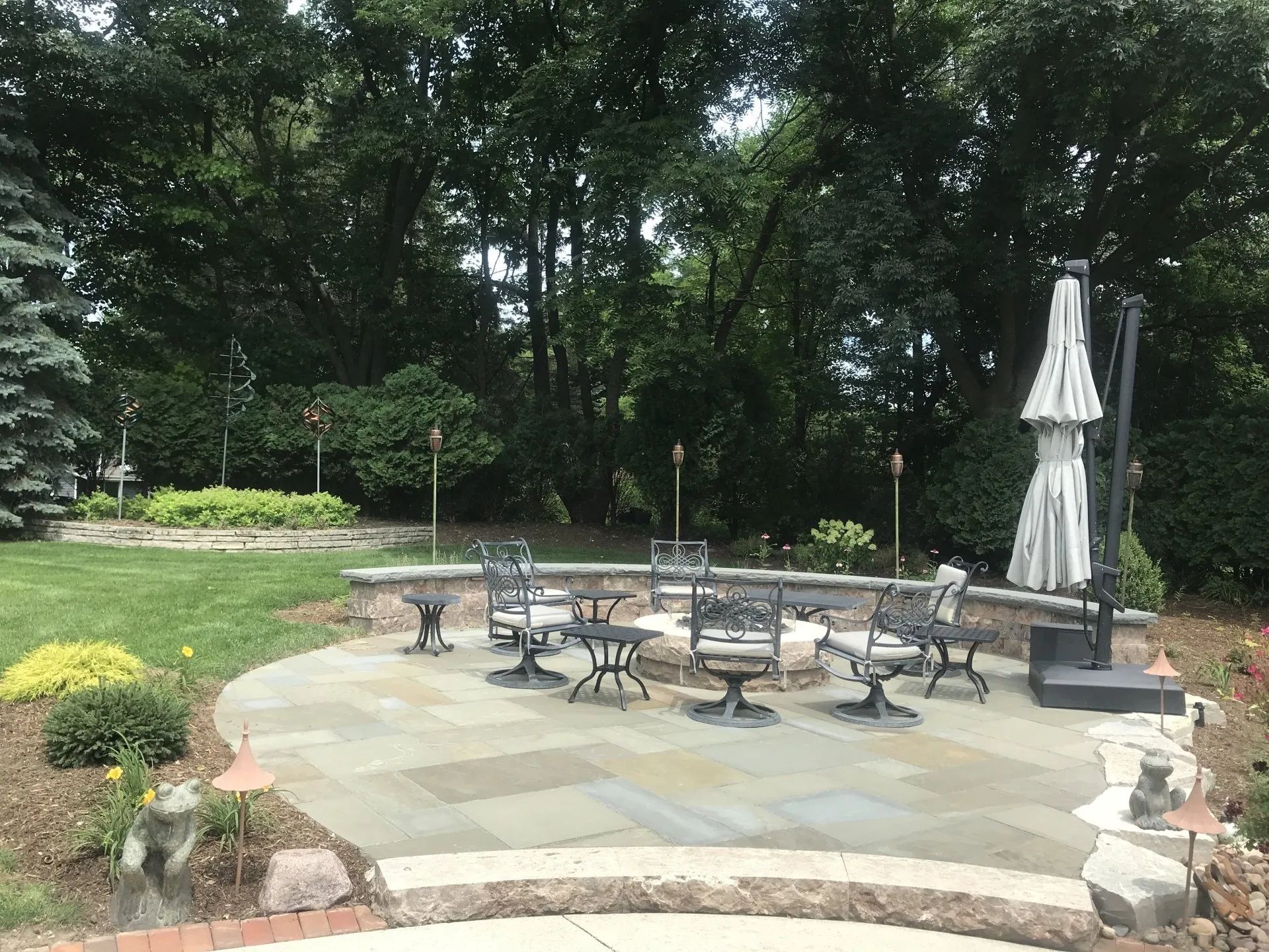 A stone patio with a central fire pit surrounded by outdoor chairs and a patio umbrella, set against a wooded backdrop.