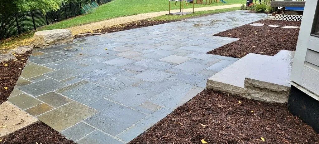A slate patio with square tiles surrounded by brown mulch, bordered by a green lawn and a stone retaining wall.