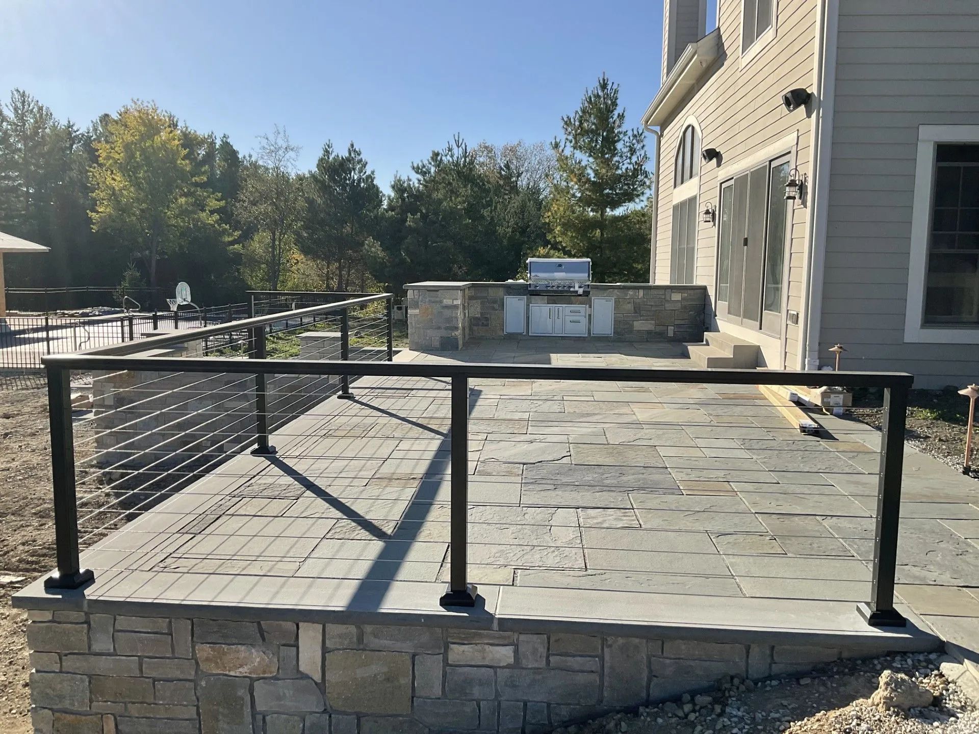 A stone patio with a built-in outdoor kitchen, featuring a black metal cable railing overlooking a backyard.