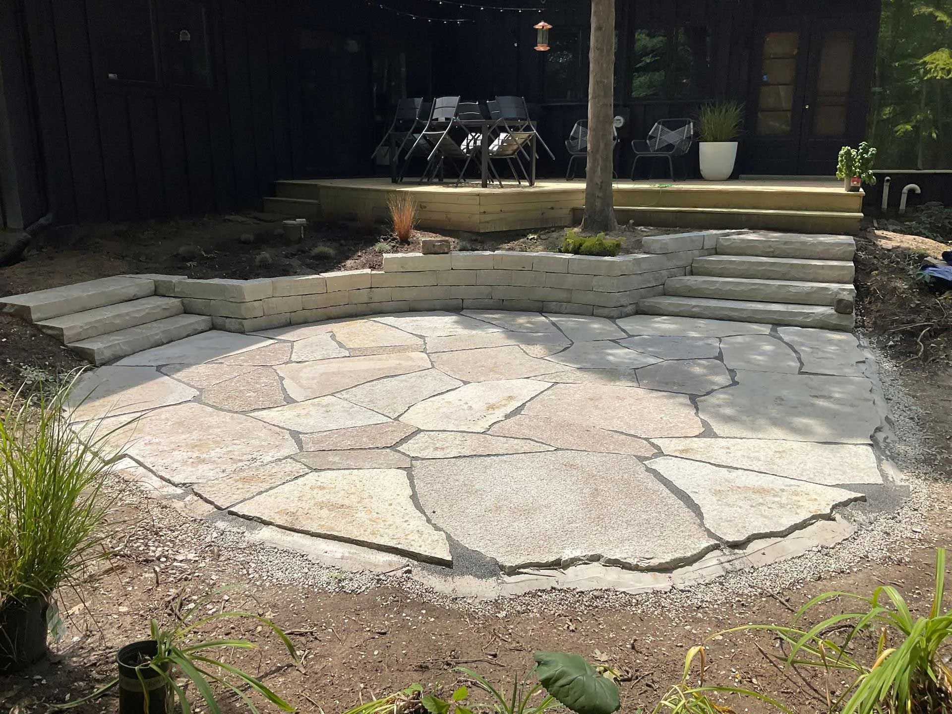 A circular stone patio features two stone staircases leading to a deck at a house with dark siding.