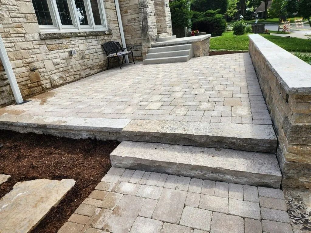 Stone patio with paver flooring, retaining wall, and steps leading to a house entrance.