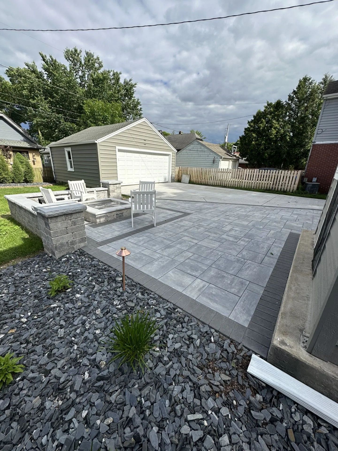A modern gray paver patio featuring a built-in fire pit with white chairs, adjacent to a detached garage under a cloudy sky.