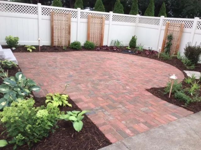 A semicircular red brick patio surrounded by a white fence, landscaped garden beds, and wooden trellis accents.