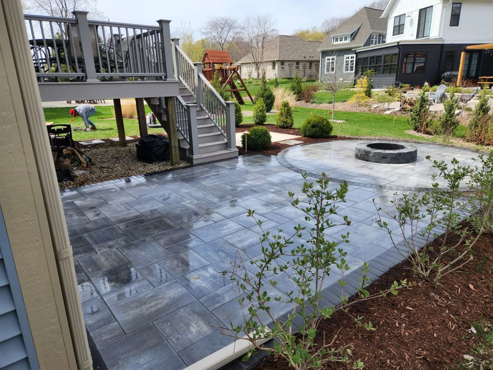 A gray stone patio featuring a circular fire pit, situated beneath a wooden deck with stairs in a grassy backyard.