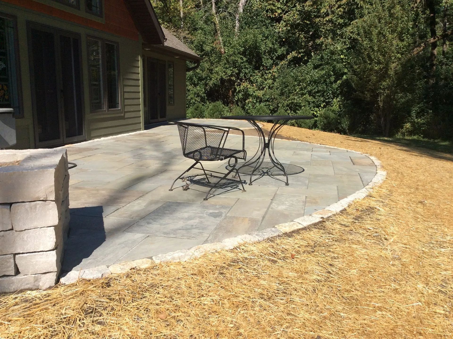 A stone patio with a metal table and chair beside a house, surrounded by fresh wood mulch.