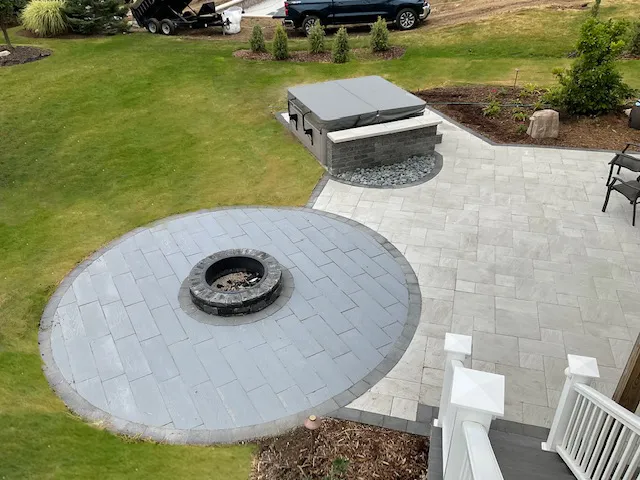 High-angle view of a backyard stone patio featuring a round fire pit area and a raised hot tub with a gray cover.