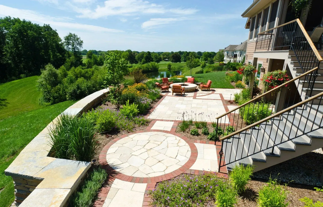 Sunny landscaped patio with stone path, circular pavers, flowers, and stairs beside a house