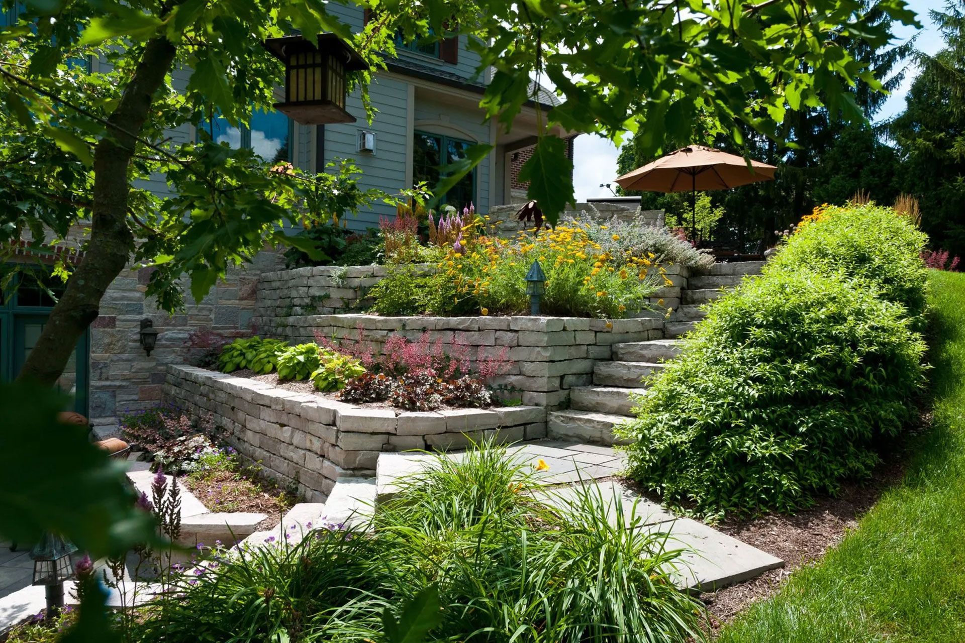 Stone tiered garden beds and staircase leading to a house patio on a sunny day.