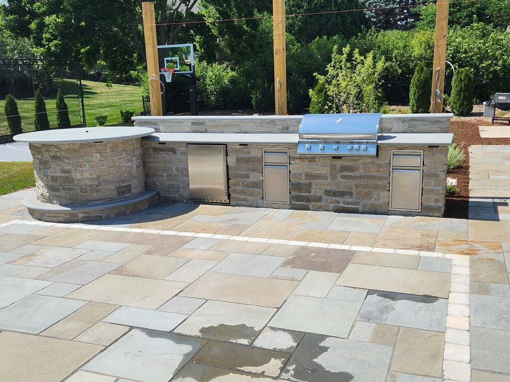 Outdoor stone kitchen with built-in grill and sink on a tiled patio, surrounded by greenery