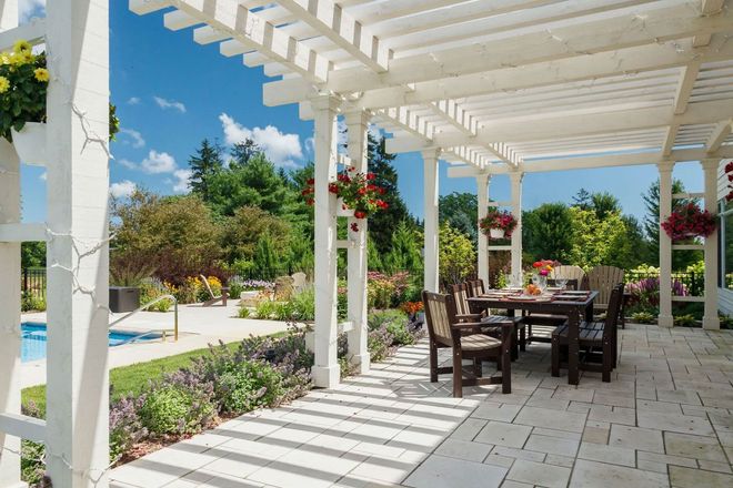 A white wooden pergola shades a stone patio with a dining table and chairs, overlooking a sunny garden and pool area.