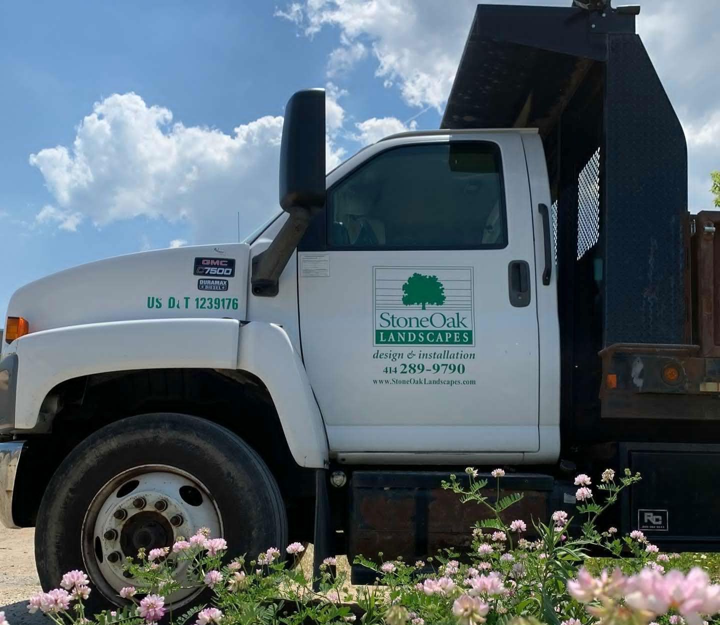 A white Stone Oak Landscapes dump truck parked outdoors, viewed from the side with flowers in the foreground.