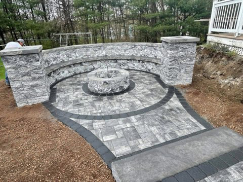 Stone patio with curved retaining wall, circular fire pit, and gray pavers in a wooded yard