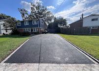 Freshly paved black driveway leading to a suburban house with green lawn and cloudy sky