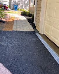 Driveway beside a beige garage, with a black asphalt foreground and a light paver walkway leading to a yard.
