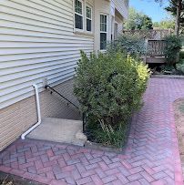 Side of house with white siding, bushes, and red brick walkway beside a downspout