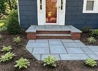 Front entry with concrete paver walkway, brick steps, and landscaped mulch beds leading to a blue house.