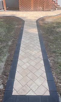 Narrow tiled walkway bordered by dark edging, leading through a grassy yard to a house entrance
