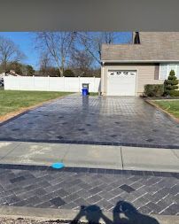 Dark gray concrete driveway leading to a garage, with a blue trash bin and white fence beside a house.