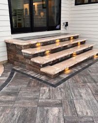 Stone front steps with wooden treads leading to a patio beside a house, with patterned pavers below