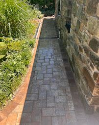 Narrow brick walkway beside a stone wall, bordered by plants and grass
