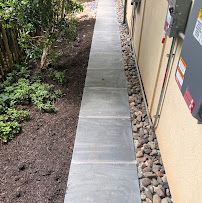 Narrow concrete path beside a building, bordered by rocks and landscaping shrubs.